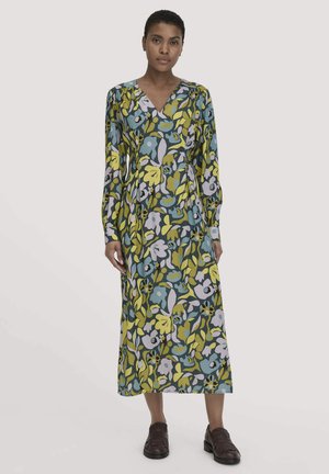 Vrouw in een midi-jurk met lange mouwen en een V-hals, voorzien van een kleurrijk bloemenpatroon, in combinatie met donkerbruine instappers, staand tegen een effen achtergrond.
