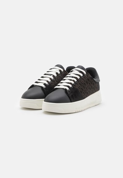 Sneaker in pelle nera con lacci bianchi e un motivo scuro testurizzato sui lati, caratterizzate da una suola bianca spessa e punta rotonda.