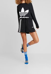 Černá mikina s bílým logem Adidas, doplněná černými šortkami s bílými bočními pruhy. Modré ponožky s bílými pruhy a černé pantofle.