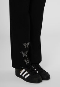 Black sweatpants with reflective butterfly designs at the hem, paired with black Adidas sneakers featuring three white stripes.