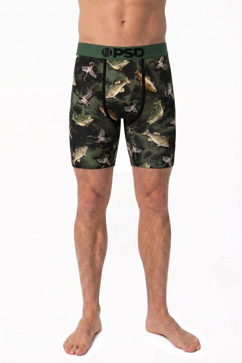 Mann trägt grüne Boxershorts mit Fisch- und Vogelmustern und steht barfuß vor weißem Hintergrund.