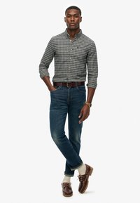 Camisa de cuadros en tonos azul marino y blanco, jeans de mezclilla azul, zapatos de barco marrones y calcetines cortos crema. El modelo posa con las manos en los bolsillos.