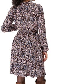 Femme portant une robe fleurie à manches longues, longueur genou, taille cintrée, associée à des collants noirs et des bottes marron, vue de dos.