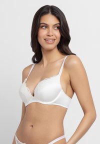 Žena s dlhými tmavými vlasmi, oblečená v bielej čipkovanej podprsenke a zodpovedajúcich nohavičkách, usmieva sa a pozerá doľava na jednoliatom pozadí.