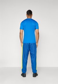 Niebieska koszulka sportowa z krótkim rękawem w połączeniu z niebieskimi spodniami dresowymi z żółtymi paskami po bokach, wykonane z gładkiego, lekkiego materiału.