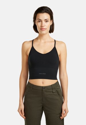 Frau mit kurzen dunklen Haaren, die ein schwarzes, bauchfreies Tanktop und olivgrüne Hosen trägt, stehend vor einem schlichten hellen Hintergrund.