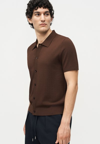 Άνδρας με μαύρα σγουρά μαλλιά που φοράει καφέ πουκάμισο με κοντά μανίκια και κουμπιά και σκούρο μπλε παντελόνι, κοιτάζει αριστερά σε ουδέτερο φόντο.