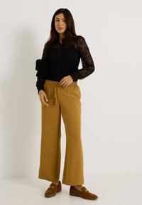 Blusa de encaje negro de manga larga con detalle de botones, combinada con pantalones anchos color coñac con cintura ajustable con cordón y adorno de borlas. Mocasines marrones.