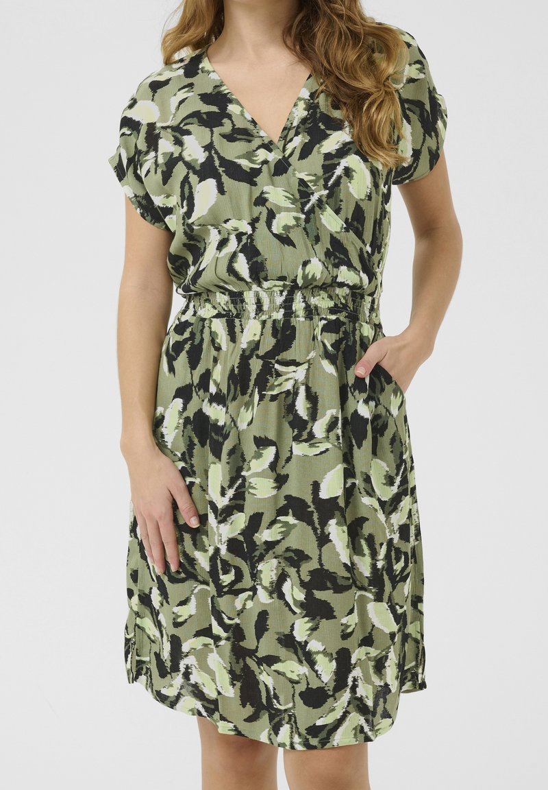 Femme portant une robe verte à manches courtes avec un motif abstrait de feuilles noir et blanc et une taille élastique, une main dans la poche.
