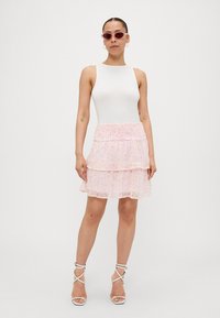 Bodysuit blanc sans manches associé à une jupe rose à couches ornée d'un motif floral, complété par des talons blancs à lanières et des lunettes de soleil.