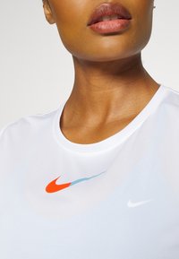Camiseta atlética blanca con cuello redondo, que presenta un gran logotipo de swoosh de Nike en rojo y azul y un swoosh más pequeño en blanco en el lado izquierdo.