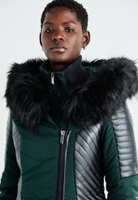 Pateikta zaļā jaka ar melnu mākslīgas kažokādas kapuci, ribotām melnām plecu detaļām un priekšējo rāvējslēdzēju. Struktūrizēts audums ar dūnu patteriem.