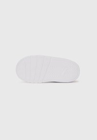Semelle de sneaker blanche avec un motif de tread texturé et un logo subtil visible près de la zone du talon sur un fond blanc uni.