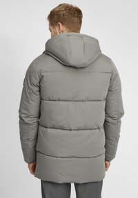Veste matelassée grise avec capuche, présentant des sections matelassées, une texture lisse et des poignets côtelés. Design minimaliste sans logos visibles.