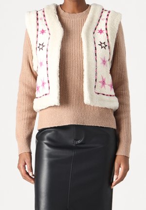 Henkilöllä on päällään beige ribbineulepusero, valkoinen fleece-liivi, jossa on vaaleanpunaisia ja ruskeita tähtikuvioita, sekä musta nahkahame.