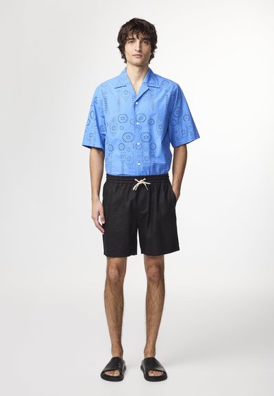 Camicia azzurra a maniche corte con motivi perforati, abbinata a pantaloni corti neri con coulisse e sandali neri scivolati. Texture liscia.