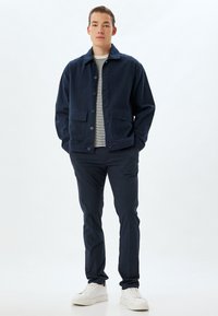 Giacca in denim blu navy con due tasche frontali, chiusura a bottoni e una camicia a righe sotto, abbinata a pantaloni scuri e sneakers bianche.