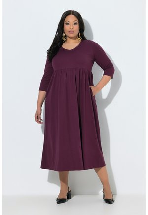 Donna in piedi su sfondo bianco che indossa un vestito midi viola con maniche a tre quarti, tacchi neri e grandi orecchini dorati.