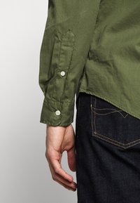 Grön skjorta med uppvikt ärm, med spetsig manschett och cremefärgade knappar, tillsammans med mörk denimjeans.