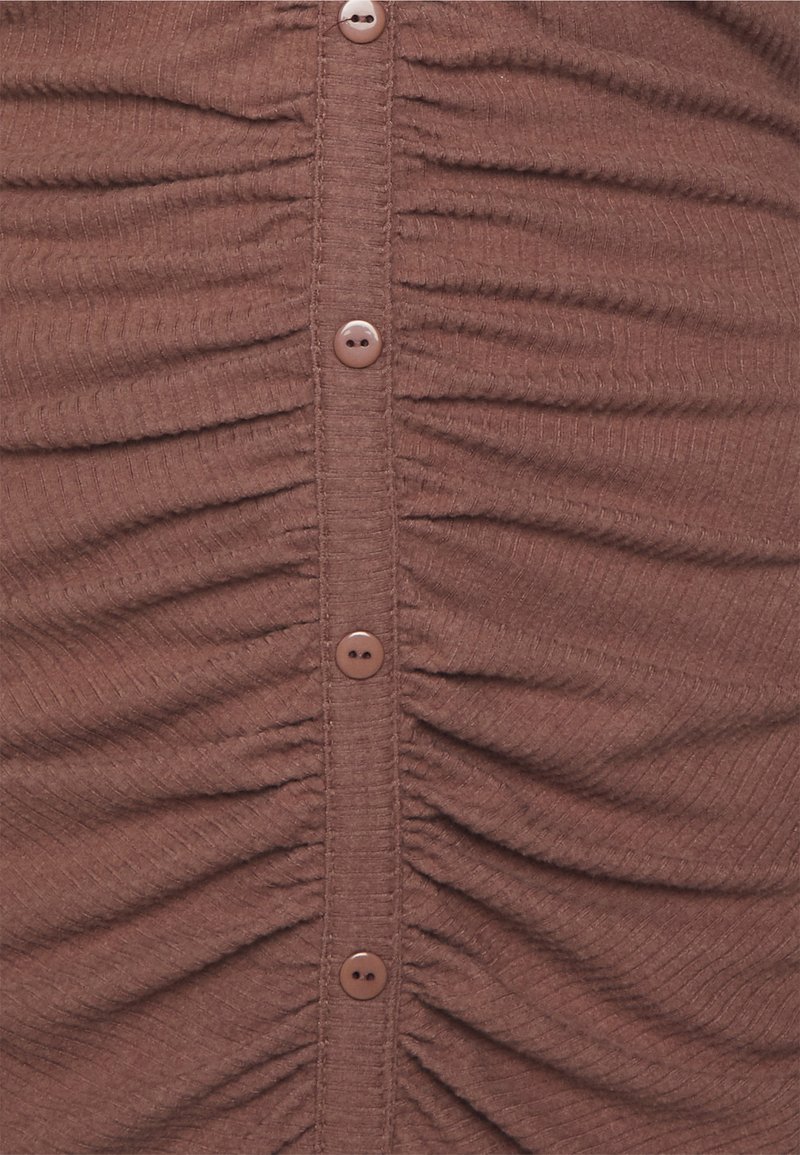 Top castanho em tecido canelado com franzido vertical e seis botões redondos na frente. Textura suave, material macio.