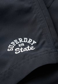 Melnas audums ar baltu izšuvumu, kas lasa "SUPERDRY ATH State." Ievērojama tīra šuvju kvalitāte un gluda tekstūra, koncentrējoties uz zīmola detaļām.