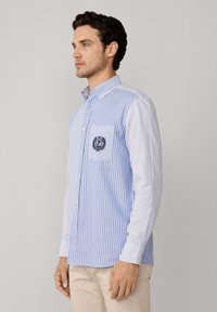 Blauw-wit gestreepte blouse met knoopsluiting, voorzien van een borstzak met een geborduurd logo, lange mouwen en een klassieke kraag.