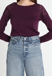 Kobieta w purpurowej bluzce z długim rękawem, ozdobionej koronką na ramionach, oraz w jasnoniebieskich dżinsach z wysokim stanem.