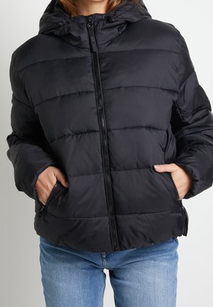 Persoon draagt een zwart doorgestikt gewatteerd jack met capuchon, voorzien van ritszakken en een ritssluiting aan de voorkant, gecombineerd met lichtblauwe spijkerbroek.