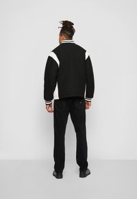 Svart varsity-jakke med hvite detaljer, ribbet krage og mansjetter, båret over svarte bukser og sko, som viser en avslappet passform og rene linjer.