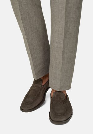 Mocassins en daim marron portés avec un pantalon sur mesure à carreaux beige, montrés de la cheville au pied sur un fond blanc.