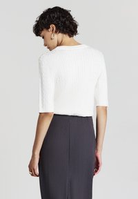 Haut blanc en tricot à manches courtes avec un motif en câble texturé, associé à une jupe ajustée grise foncé. Le haut présente un col rond.