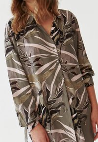 Donna che indossa una camicia a maniche lunghe con bottoni, caratterizzata da un motivo astratto di foglie e righe in tonalità di verde tenue, beige, marrone e nero.