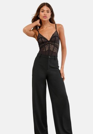 Schwarzer transparenter Bodysuit mit Spitze, der über einen tiefen Ausschnitt und dünne Träger verfügt, kombiniert mit hoch taillierten schwarzen weit geschnittenen Hosen.