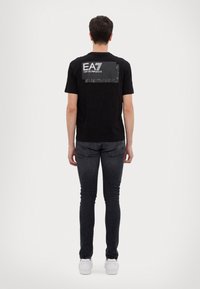 Juodas medvilninis marškinėlis su kamufliažo detalėmis ir "EA7 Emporio Armani" logotipu gale. Liemenukas, trumpomis rankovėmis ir apvalia kaklo iškirpte.