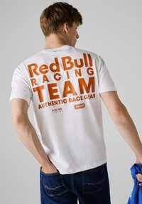 Bijela majica kratkih rukava s velikim narančastim tekstom na stražnjoj strani: "Red Bull Racing TEAM AUTHENTIC RACE GEAR," s logotipima i jednostavnim dizajnom.