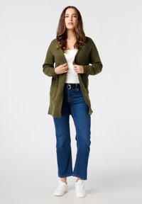 Olijfgroene cardigan met een open voorkant, lange mouwen en zakken, gecombineerd met een witte top, blauwe jeans en witte sneakers.