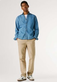 Camicia di denim con bottoni in azzurro chiaro, abbinata a pantaloni beige. Sneakers bianche con dettagli navy completano l'outfit.