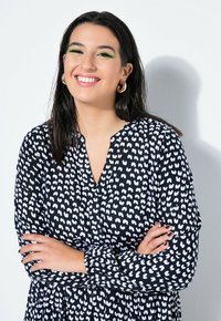 Musta mekko, jossa valkoinen sydänkuvio, pitkät hihat ja napitettu pääntie. Pitkät tummahiuksinen malli hymyilee ja käyttää kultaisia rengaskorvakoruja.