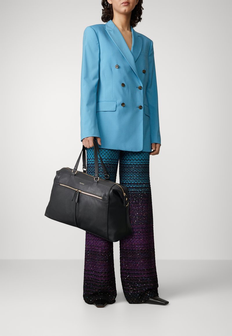 Personne portant un blazer croisé bleu vif et un pantalon large à motifs violet-bleu, tenant un grand sac de voyage en cuir noir.