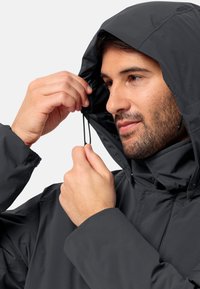 Giacca nera impermeabile con cappuccio regolabile. Caratteristiche includono cordini elastici e una consistenza morbida e liscia. Nessun motivo o ferramenta visibile.