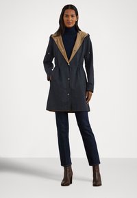 Žena v tmavě modrém kabátu s hnědým lemováním kapuce, tmavém roláku, tmavých džínách a hnědých botách na podpatku, stojící s rukama v kapsách.