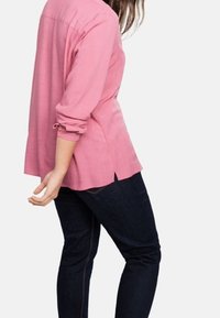 Camicia rosa a maniche lunghe con colletto e spacchi laterali, abbinata a jeans blu scuro. Tessuto morbido, vestibilità rilassata e maniche arrotolate.