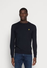 Maglione a girocollo blu navy in maglia con maniche lunghe, con un piccolo emblema giallo sul petto. Indossato con jeans blu.