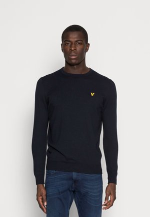 Maglione a girocollo blu navy in maglia con maniche lunghe, con un piccolo emblema giallo sul petto. Indossato con jeans blu.