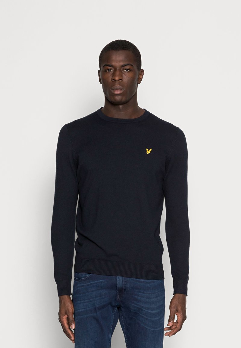 Maglione a girocollo blu navy in maglia con maniche lunghe, con un piccolo emblema giallo sul petto. Indossato con jeans blu.