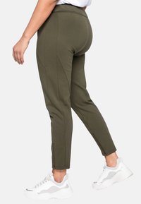 Pantaloni tapered verde oliva con pannelli laterali e zip alle caviglie. Il tessuto appare liscio ed elasticizzato. Abbinati a sneakers bianche.