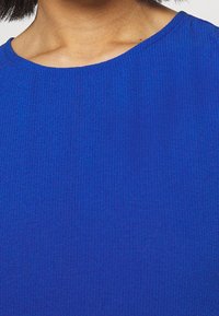 Top lavorato a maglia blu con scollo a girocollo, texture liscia e motivo a coste, incentrato sulla parte superiore del capo.