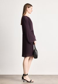 Fialové plisované šaty s dlhými rukávmi v tvare zvona, s úzkym pásom, skombinované s čiernou kabelkou a čiernymi sandálmi na podpätku. Hladká textúra látky.