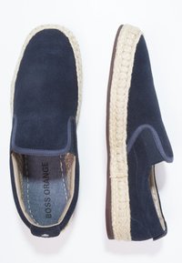 Marinblåa slip-on skor i mocka med repdetaljer runt sulan; har en vadderad innersula och en kontrasterande brun gummisula.