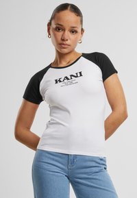 Balts piegriezts T-krekls ar melniem raglanu piedurknēm. Uz krūšu ir centrēts "KANI" logos melnā krāsā. Mīksts kokvilnas audums, vienkāršs dizains.